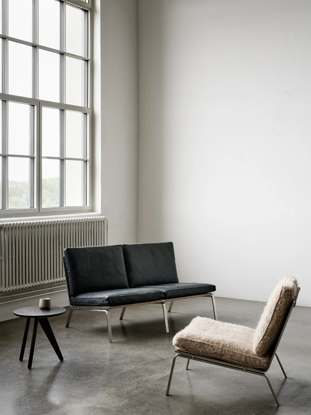 NORR11 Man 2 Seater Sofa, a sleek modern minimalist sofa featuring deep black leather upholstery and a sturdy brushed steel frame with a visible natural canvas sling base. This low-profile, industrial chic lounge sofa offers clean lines and a contemporary aesthetic. Perfect for modern living rooms, compact apartment spaces, or stylish office lounge areas.