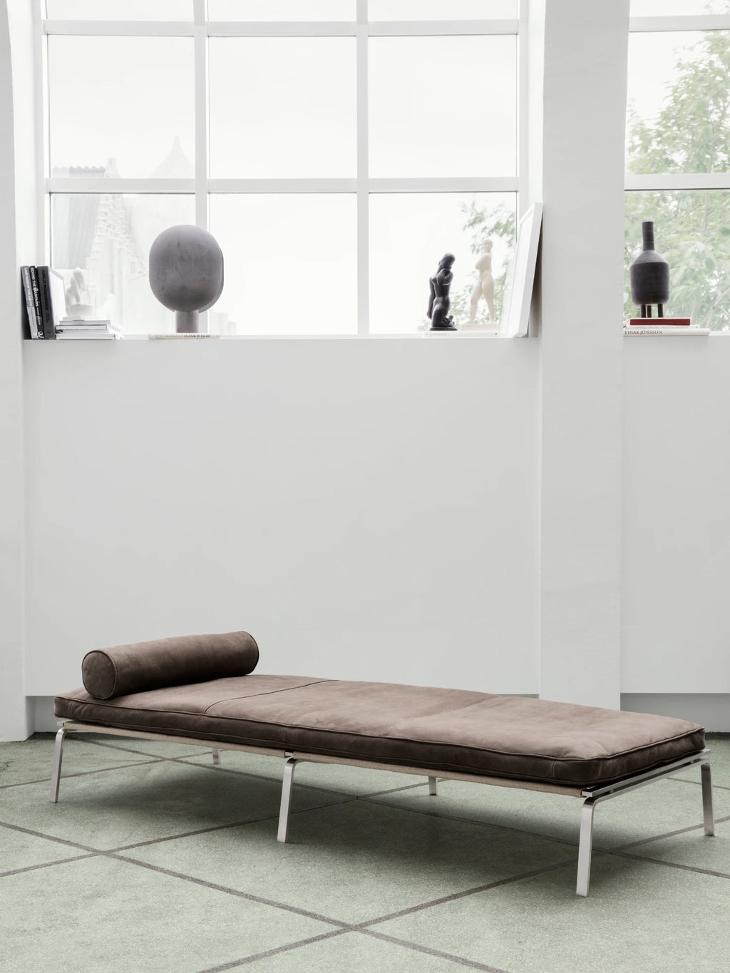 NORR11 Man Daybed, a modern minimalist daybed featuring rich brown aniline leather upholstery with a matching cylindrical bolster pillow, supported by a sleek brushed steel frame with clean, architectural lines.