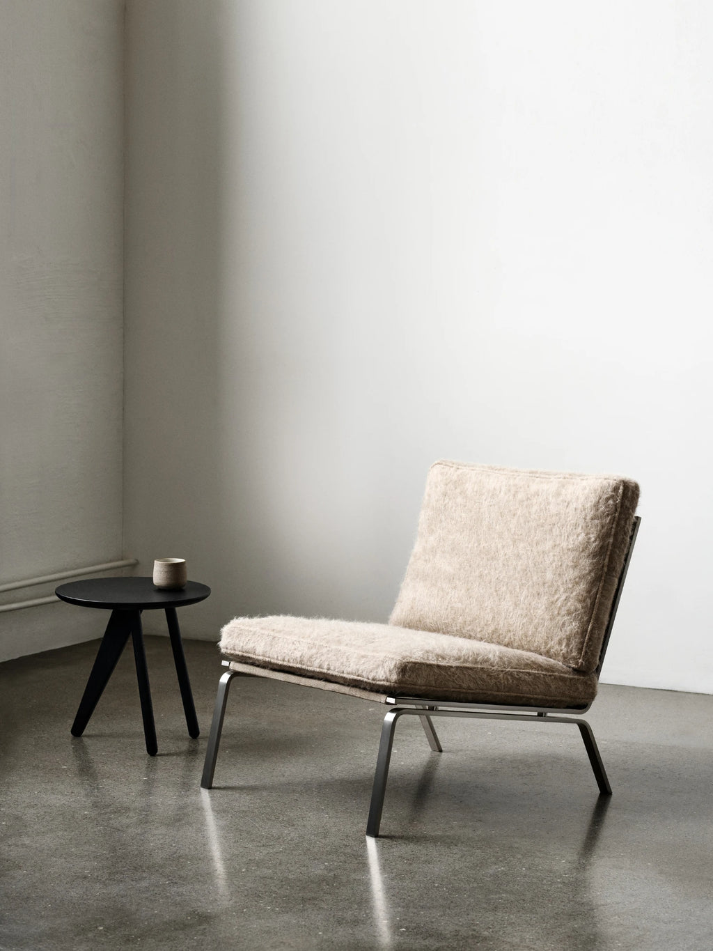 NORR11 Man Lounge Chair, a modern minimalist lounge chair featuring plush, shaggy beige upholstery, likely a wool or bouclé blend, set on a sleek brushed metal frame with a low-profile silhouette. Its inviting texture and contemporary design make it a perfect Scandinavian accent chair for a stylish living room, bedroom, or reading nook, complemented by a matte black round side table.