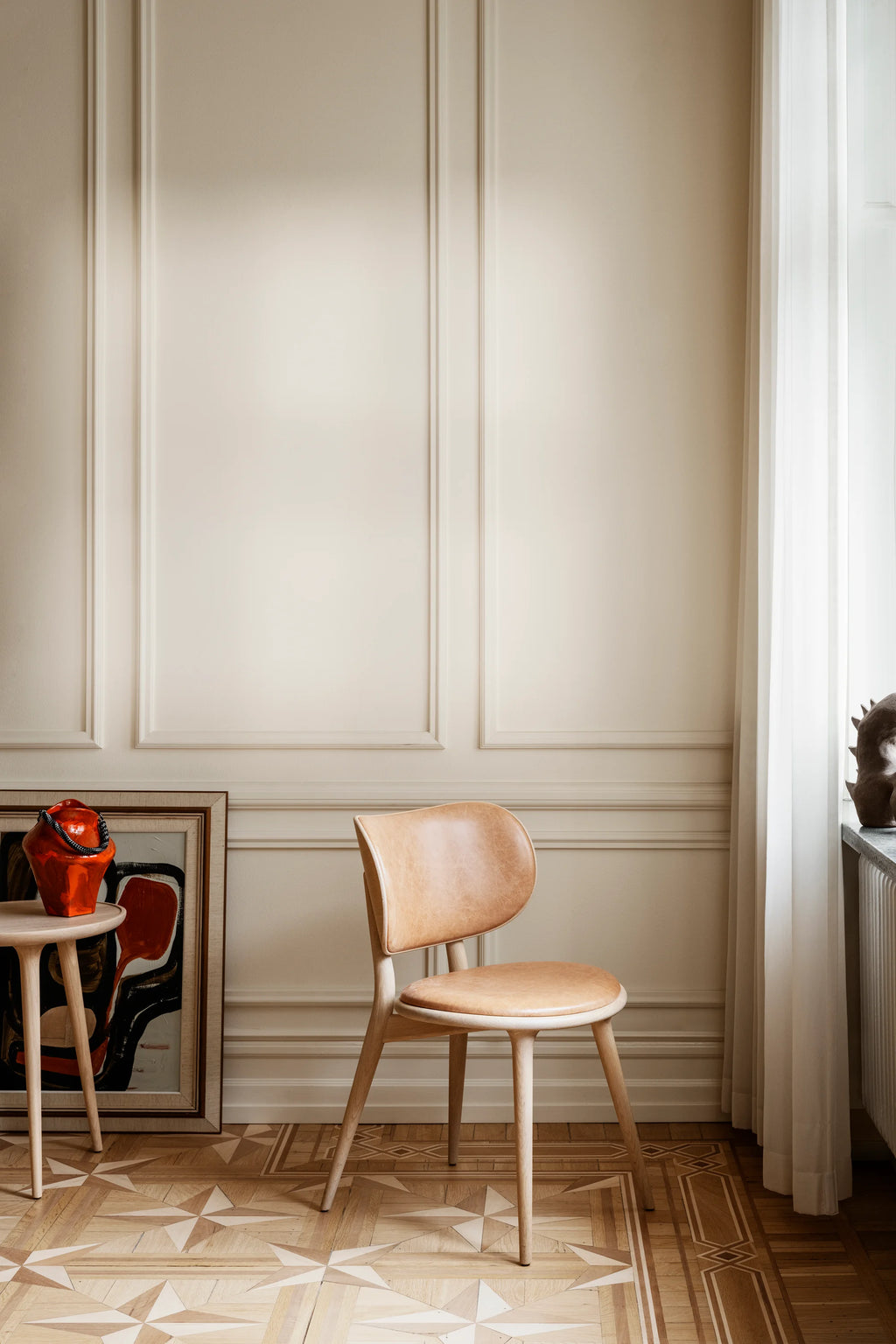 Mater The Dining Chair in light matt lacquered oak with a tan leather seat, standing elegantly against a classic cream paneled wall. The chair's minimalist design and natural wood grain are prominent, offering a timeless aesthetic.