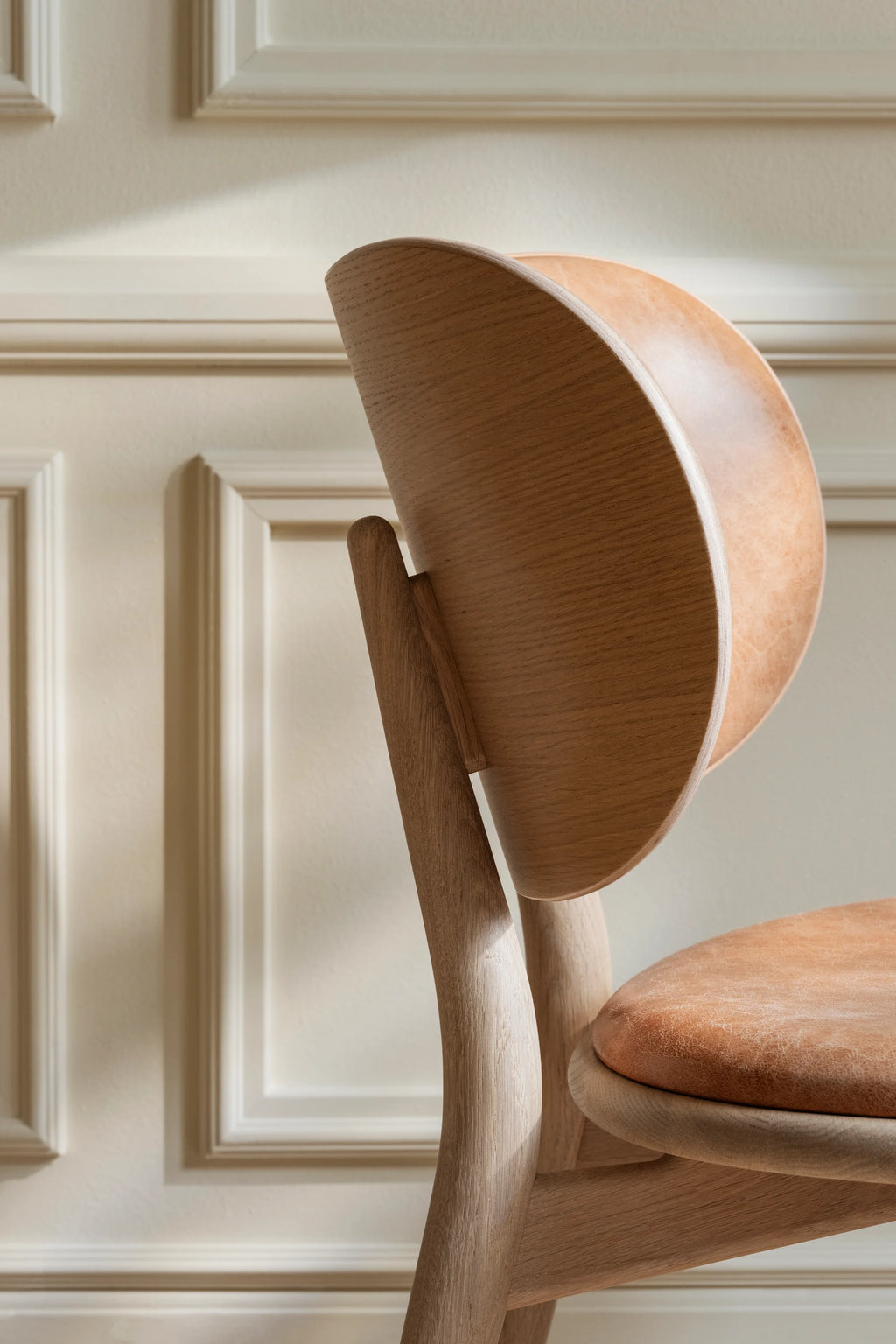 Detailed view of Mater The Dining Chair, showcasing its curved backrest in light matt lacquered oak and a textured tan leather seat. The craftsmanship and natural materials are beautifully highlighted against a classic paneled wall.
