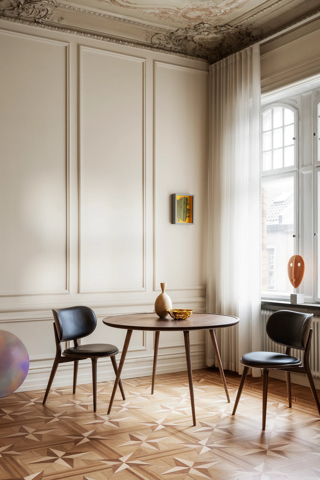 Two Mater The Dining Chairs in dark stained wood with black leather seats, flanking a matching dark wood round dining table. The chairs feature a sleek, modern design, complementing the elegant room with ornate ceilings and patterned wooden floors.