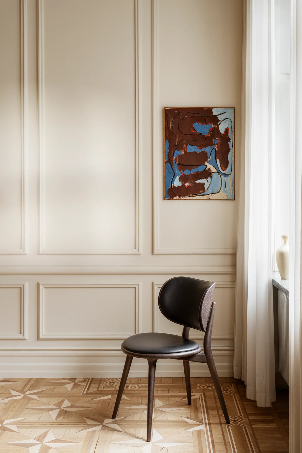Mater The Dining Chair in Sirka Grey Stained Oak with a black leather seat, standing against a sophisticated cream paneled wall. The chair's dark, rich wood finish and elegant curved backrest create a striking, modern contrast in the room.