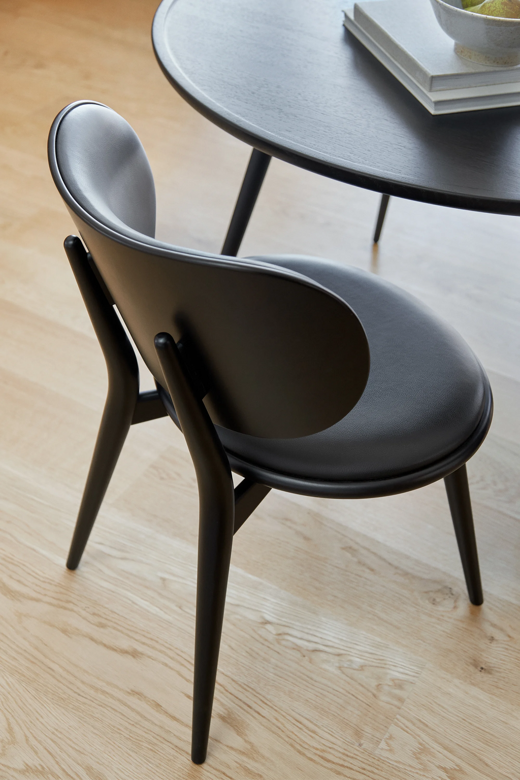Mater The Dining Chair, an elegant black dining chair with a curved backrest and padded seat, featuring sleek black wooden legs, positioned next to a matching black accent table on a light wooden floor, showcasing its modern design.