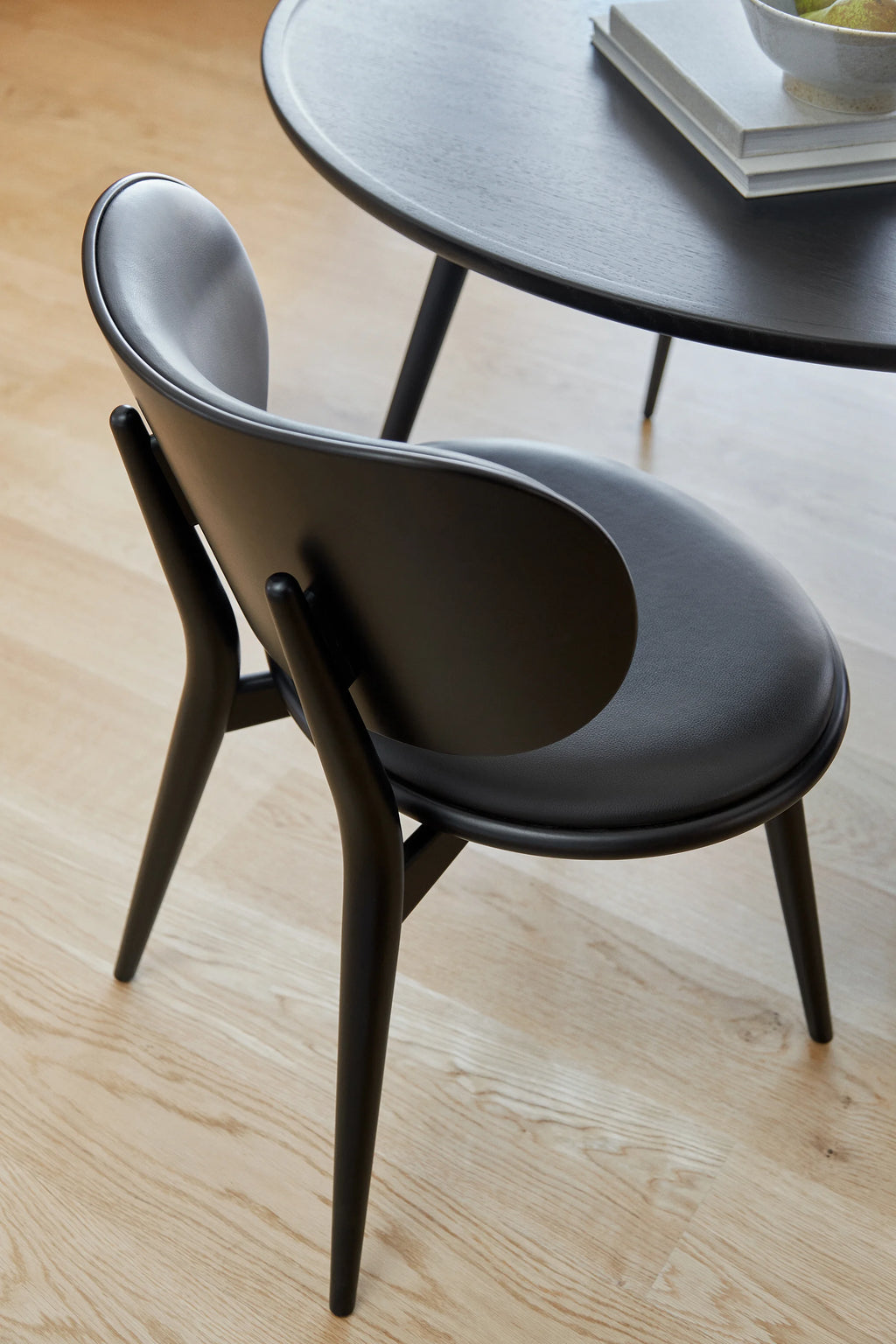 Mater Accent Round Dining Table, showcasing a close-up of a dark stained oak table and a matching dining chair with a sleek, curved backrest and black upholstered seat, highlighting the premium craftsmanship on a light wooden floor.