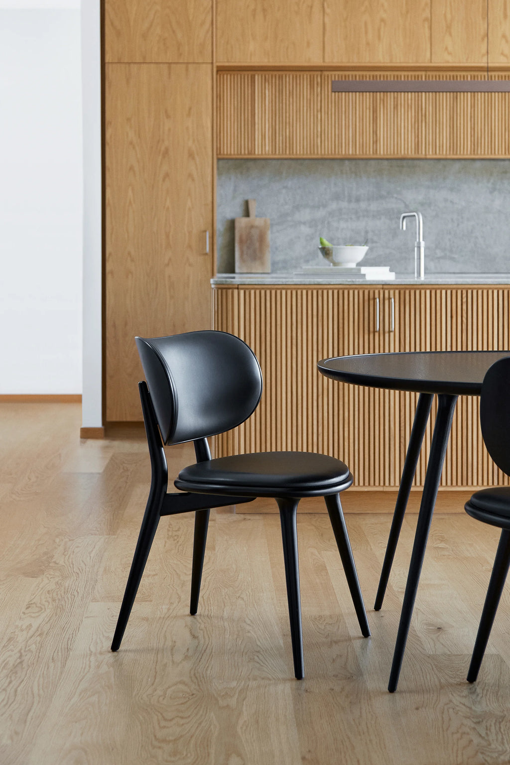 Mater The Dining Chair, a stylish black dining chair with a gracefully curved back and comfortable padded seat, paired with a black accent table, set in a contemporary kitchen with light wooden cabinetry and a marble countertop.