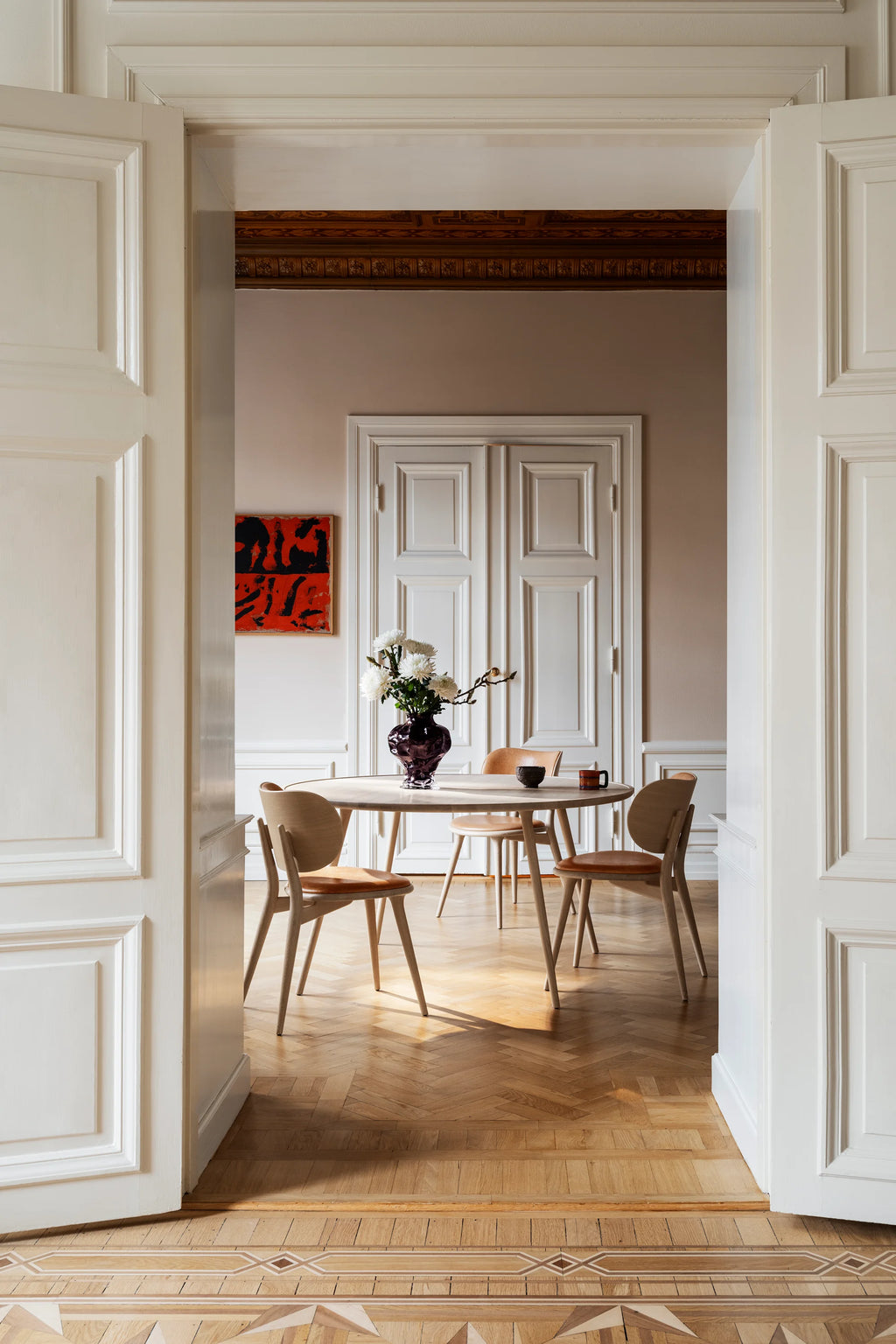 Mater Accent Round Dining Table, a light oak finish, surrounded by matching wooden dining chairs with light fabric seats, elegantly placed in a spacious, classic dining room with white paneled walls and hardwood floors.