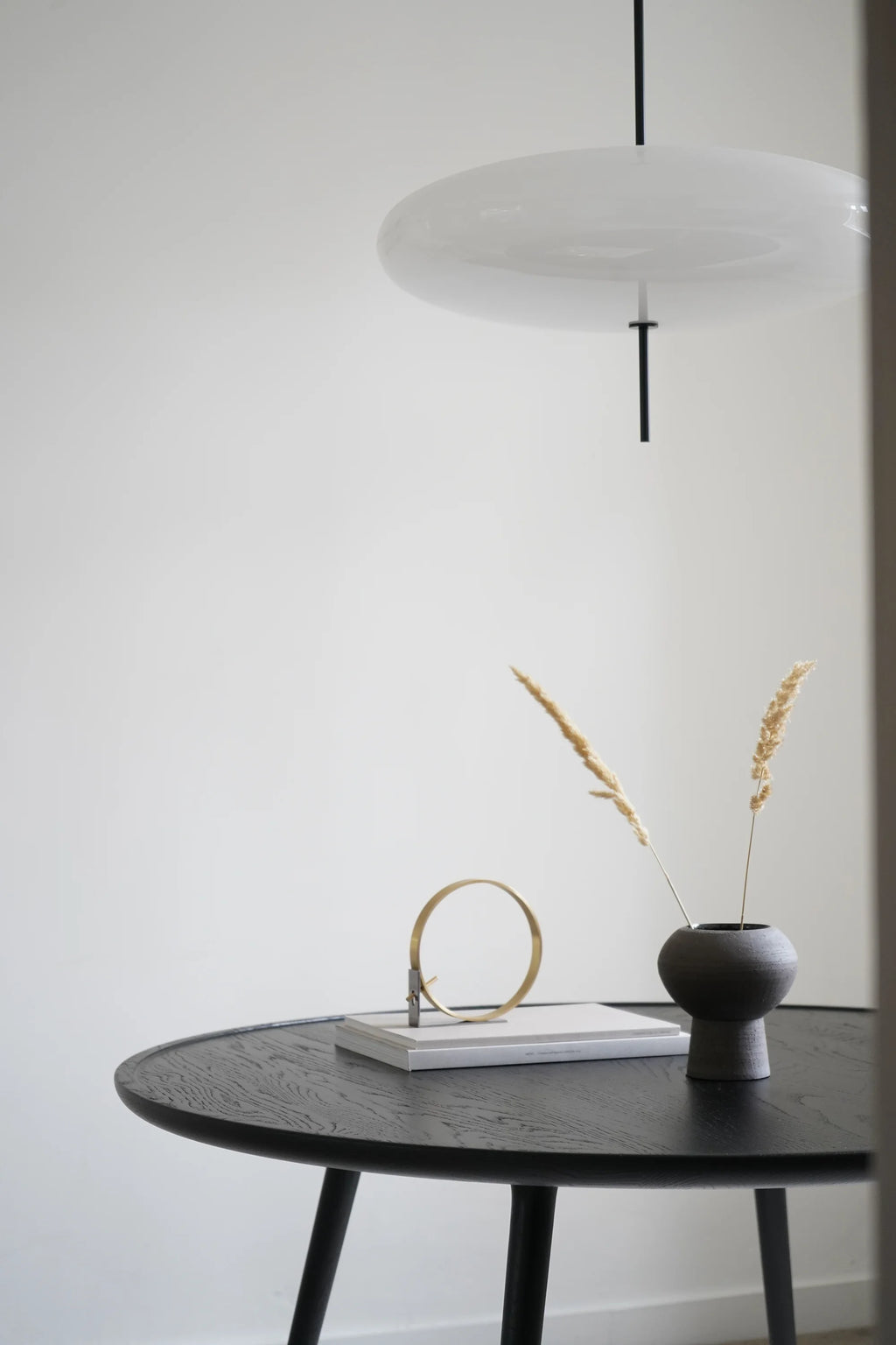 Mater Accent Round Dining Table in a dark, elegant wood finish, styled in a contemporary setting with a modern pendant light, a small vase with dried grasses, and decorative objects, creating a chic and inviting atmosphere.