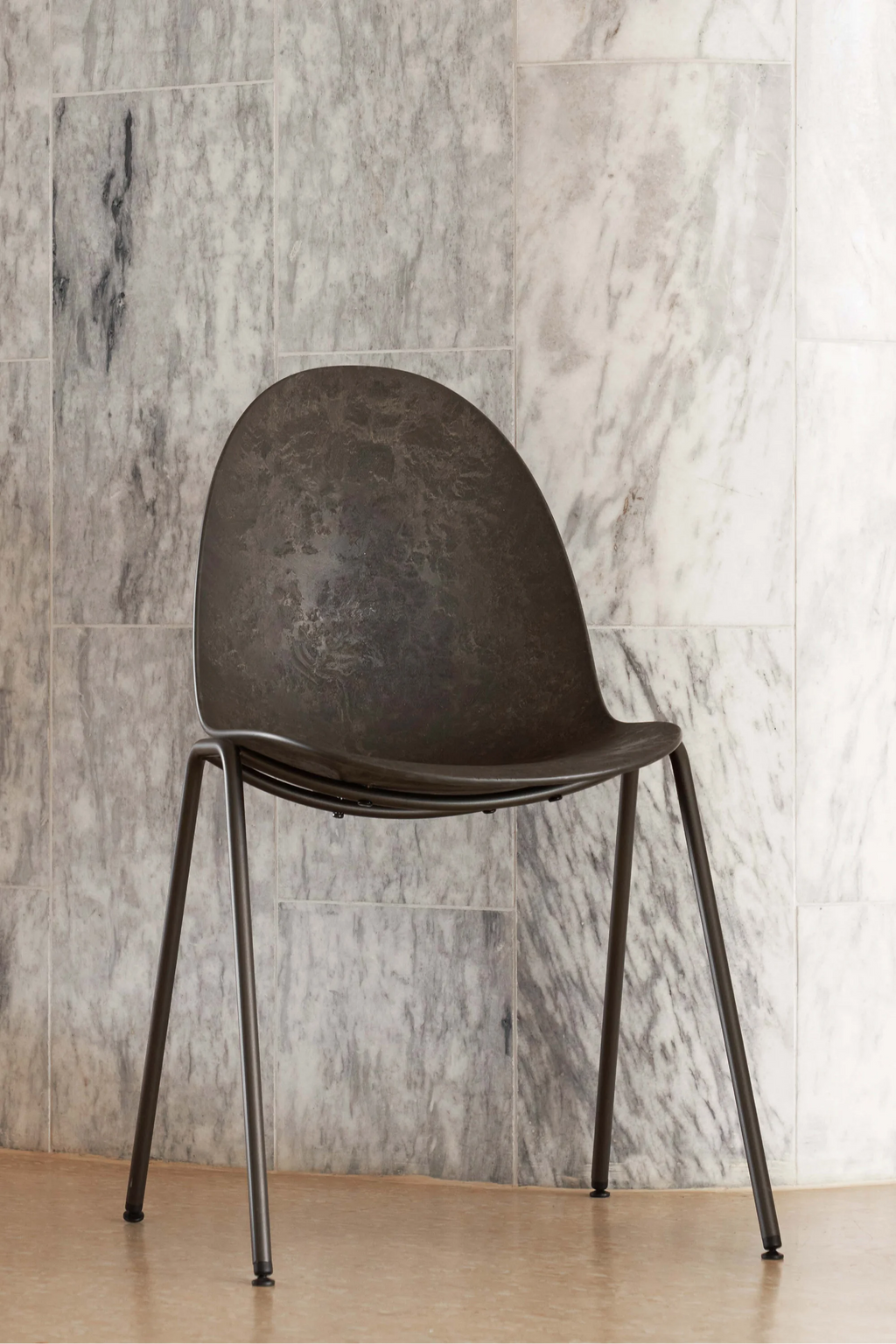 Mater Eternity Sidechair, a contemporary design with a dark, mottled gray seat and slender black metal legs, standing against a stylish gray marble tiled wall. The chair's unique texture and minimalist aesthetic are perfect for modern interiors.
