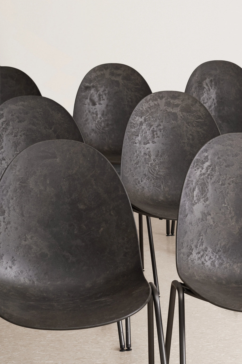 Mater Eternity Sidechair, a collection of several dark gray chairs arranged in a row, viewed from a low angle. Each chair features a subtly textured, rounded backrest and seat, supported by slender black metal legs. The chairs are positioned against a light, neutral wall, emphasizing their modern, minimalist design and uniform appearance.