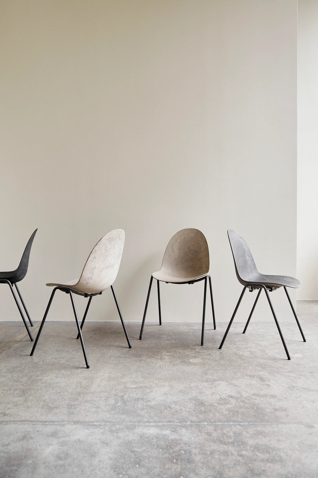 Mater Eternity Sidechairs in various earthy tones (black, light grey, beige, dark grey) lined up against a minimalist light beige wall, showcasing the chairs' versatile and modern design in a group setting.