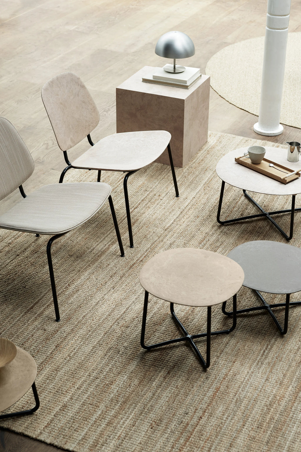 Mater Compound Lounge Chair in a contemporary setting, showcasing its light beige upholstered seat and backrest with a subtle textured pattern. The chair's minimalist black metal frame is visible, positioned on a natural woven rug. Several small round side tables and a square pedestal table complete the inviting, modern arrangement.