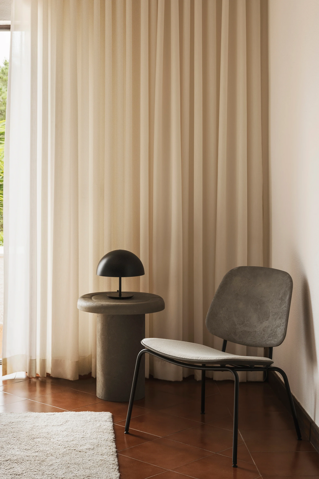 Mater Compound Lounge Chair in a modern living space. This elegant chair features a light gray upholstered seat and backrest, supported by sleek black metal legs. A black dome lamp sits on a concrete side table, all against a backdrop of flowing cream curtains and warm terracotta floor tiles.