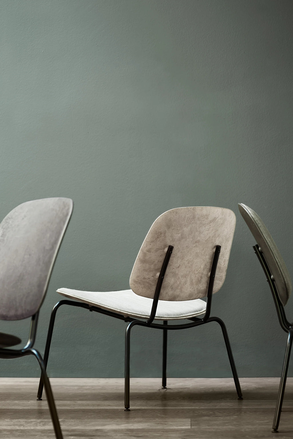 Mater Compound Lounge Chair, seen from a low angle, highlighting its unique design. The chair features a light beige upholstered seat and backrest with a subtle texture, supported by a sleek black metal frame. Two other chairs are partially visible, all set against a muted green wall and light wooden floor, creating a serene, modern aesthetic.