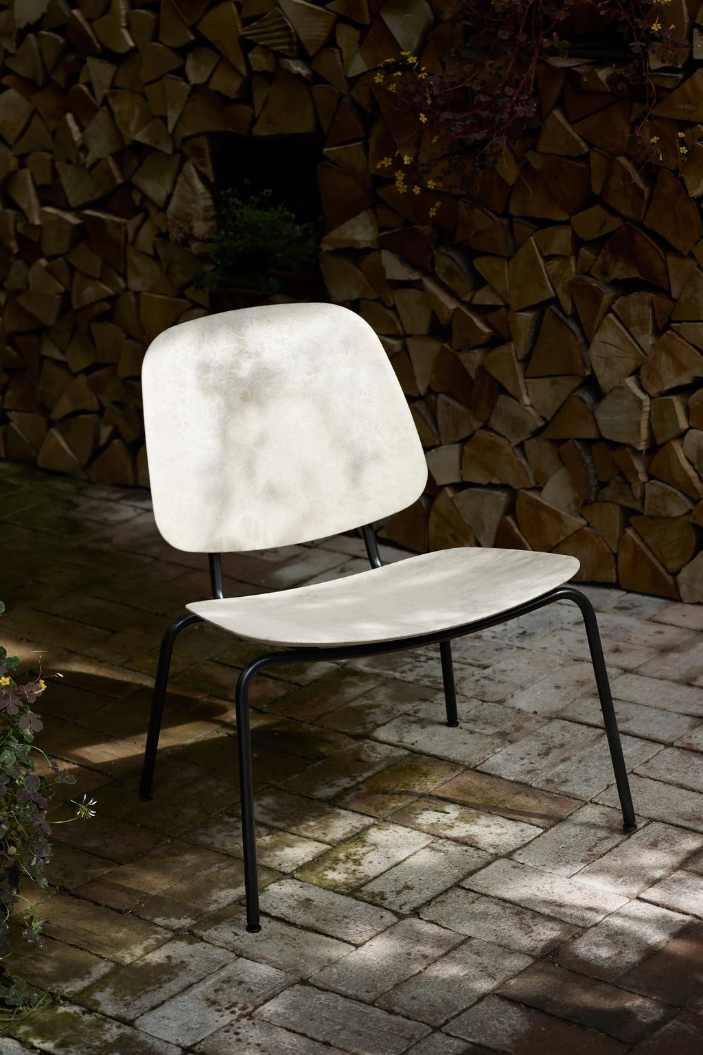 Mater Compound Lounge Chair, featuring a light, natural-toned seat and backrest with a subtle textured pattern, supported by sleek black metal legs, set on a brick patio next to a stacked woodpile.