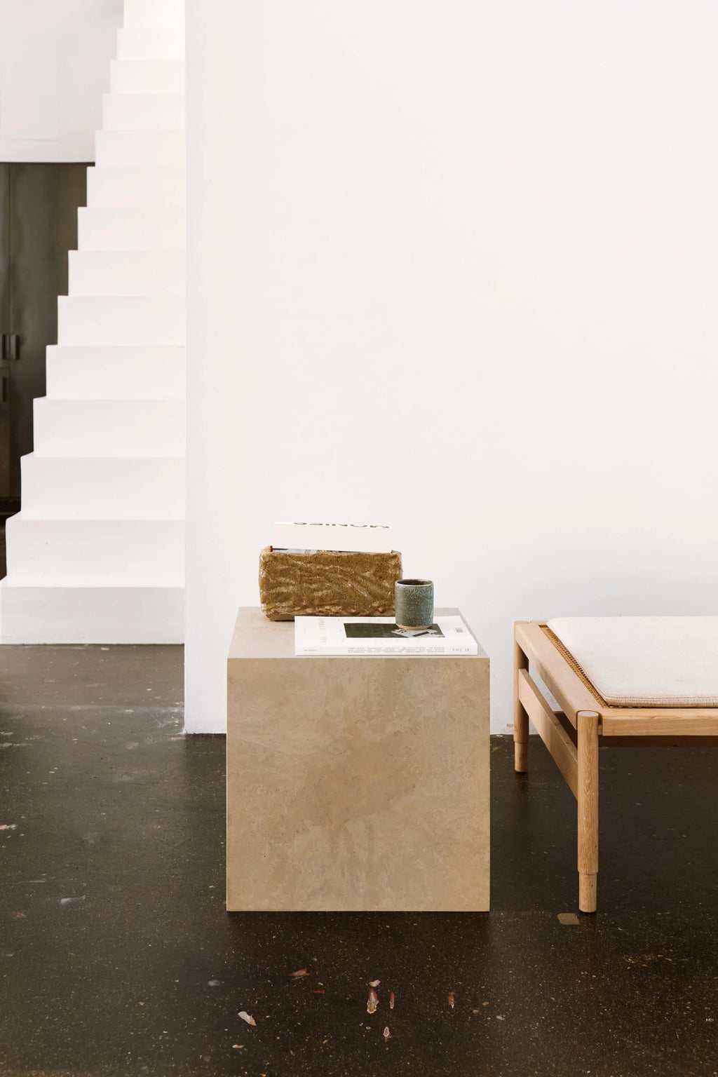 Mater Mater Cube, a minimalist and modern side table or stool crafted from light beige wood waste material, showcasing a subtle textured finish, positioned next to a wooden bench in a contemporary interior with white stairs.