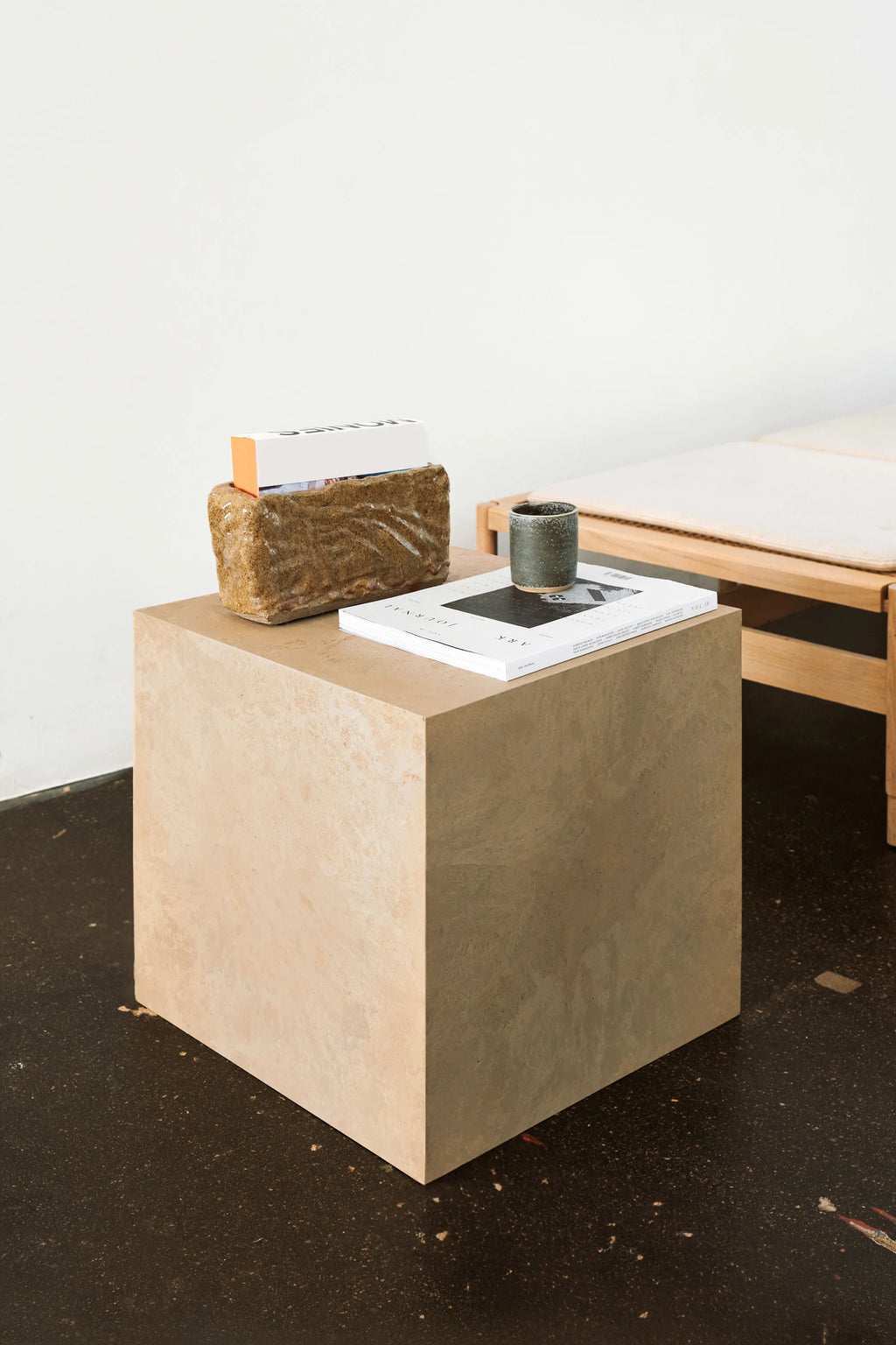 Mater Cube, a single light beige cube-shaped side table, featuring a natural, textured finish, adorned with books and a ceramic cup, in a contemporary, minimalist living space.