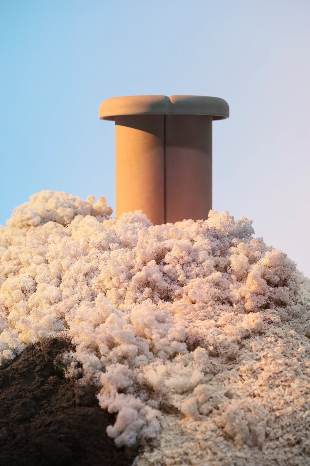 Mater Alder Stool in a warm terracotta hue, featuring a unique cylindrical base and a subtly curved top, emerging from a textured, organic-looking landscape of white and brown granular material under a soft blue sky.