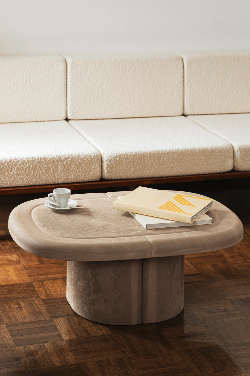 Mater Alder Coffee Table, an elegant oval-shaped design in a soft beige hue, featuring a plush, textured surface and a substantial cylindrical base, positioned on a wooden floor in a living room.