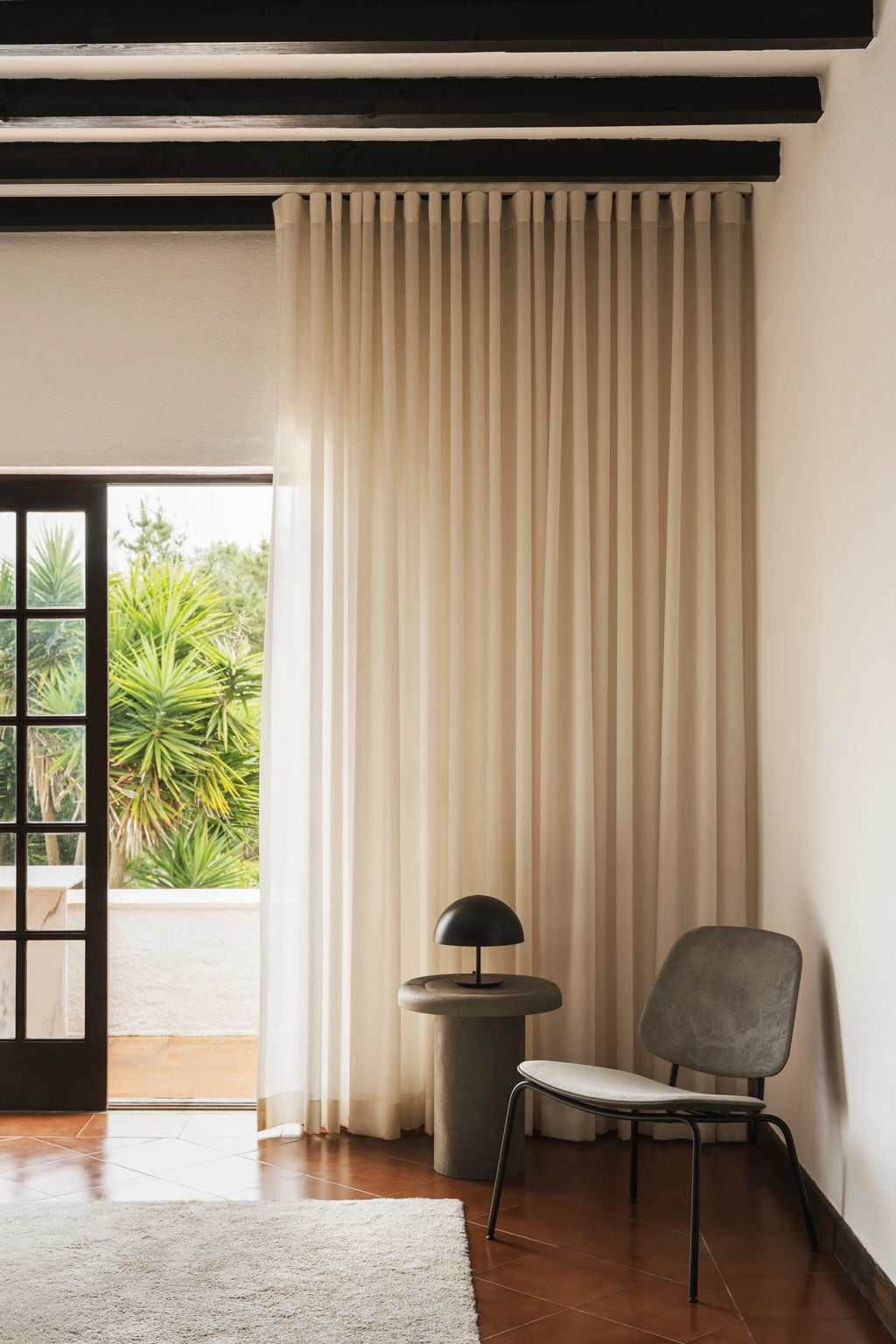 Mater Alder Side Table in a natural, earthy tone, featuring a round top and cylindrical base, positioned next to a modern lounge chair in a sunlit room with large windows and green foliage outside.