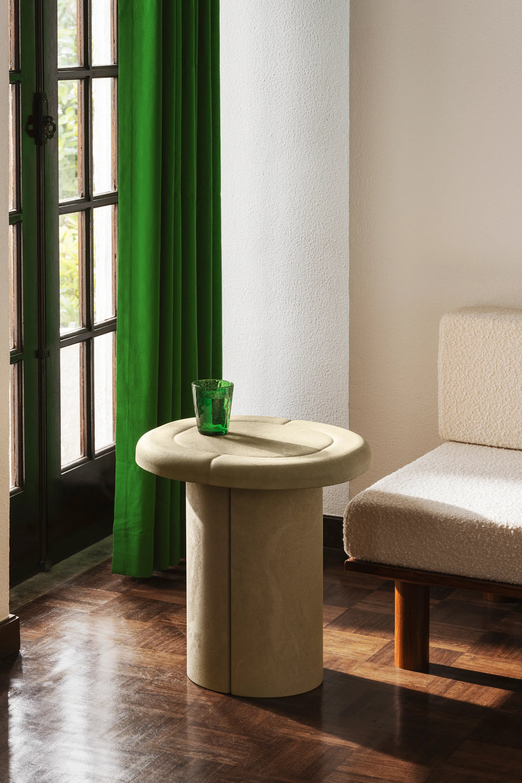 Mater Alder Side Table in a light, natural beige, featuring a round, subtly textured top and a sturdy cylindrical base, placed beside a plush, cream-colored sofa in a well-lit room with green curtains.