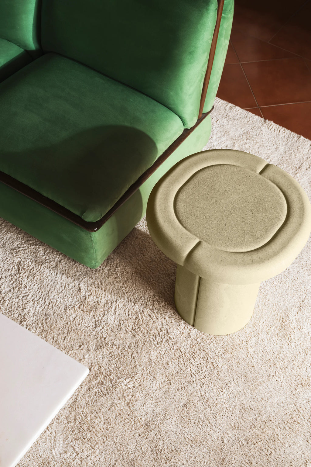 Mater Alder Side Table, an overhead shot highlighting its unique round top with subtle indentations and a robust cylindrical base, in a light beige color, positioned on a soft, textured cream rug next to a green sofa.