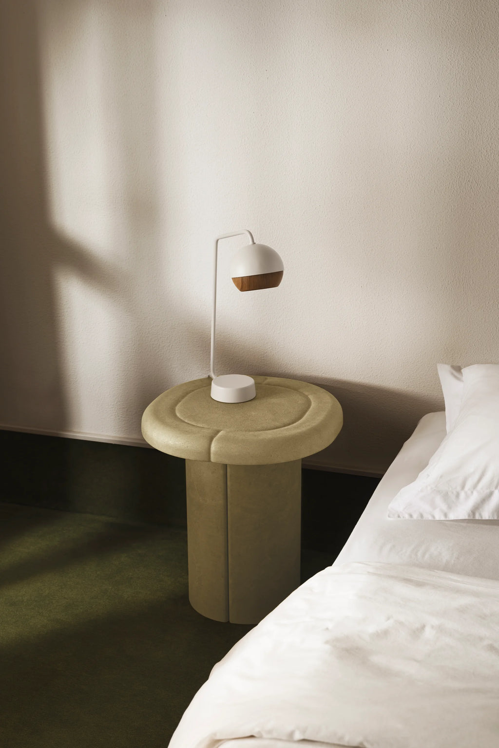 Mater Alder Side Table in a soft, light green hue, featuring a unique rounded top with subtle indentations and a sturdy cylindrical base, placed beside a bed with white linens in a modern bedroom.