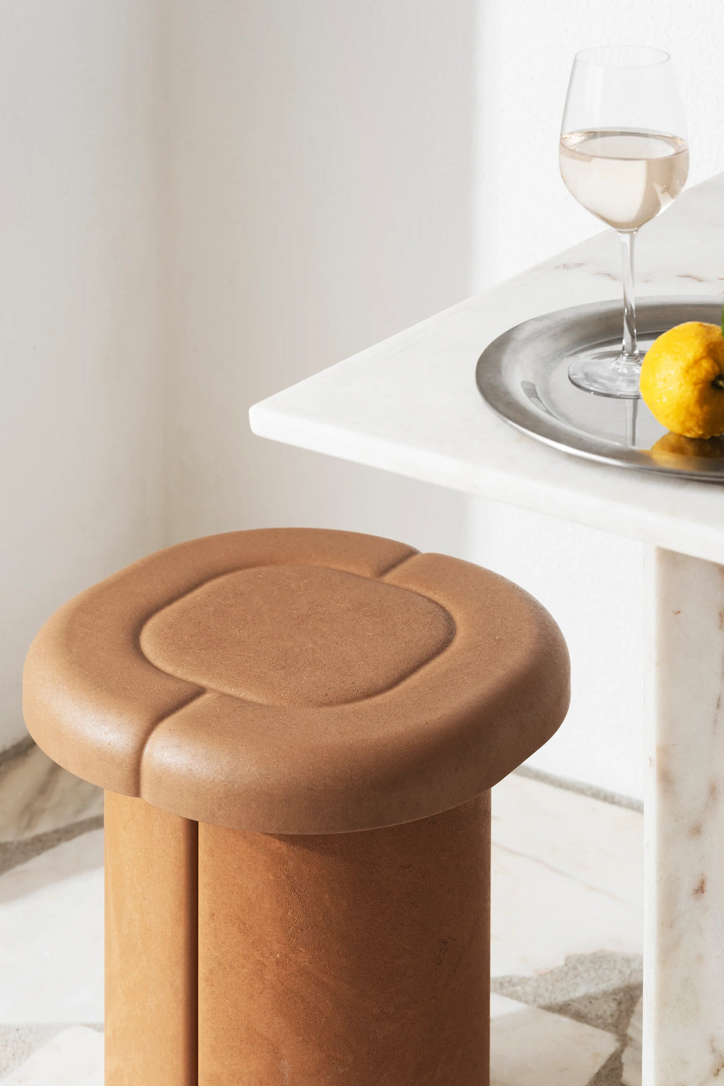 Mater Alder Stool in a warm terracotta color, showcasing its unique rounded top with subtle indentations and a sturdy cylindrical base, placed next to a white marble table with a glass of wine.