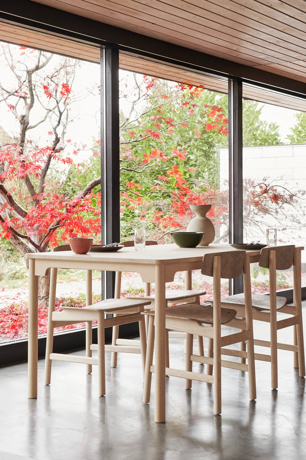 Four Mater Conscious Chairs in light, natural wood, surrounding a matching rectangular dining table in a modern dining room. The chairs feature a simple, elegant design, set against a backdrop of large windows with vibrant red autumn trees.