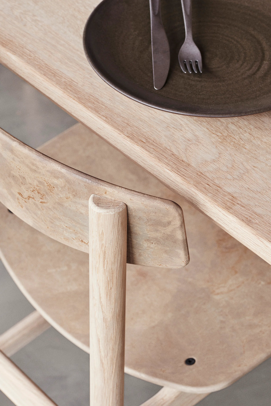 Mater Conscious Chair, a light natural wood chair with a rounded backrest, paired with a light wooden dining table. A dark brown ceramic plate with cutlery is visible, highlighting the chair's elegant, minimalist design.