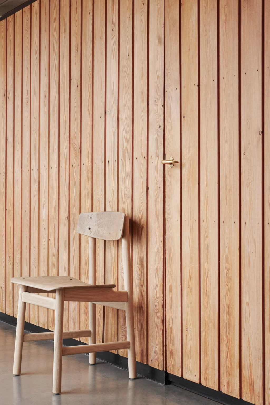Mater Conscious Chair in natural light oak, showcasing its simple yet elegant design, positioned against a warm, vertically paneled wooden wall. The chair's sturdy construction and smooth finish highlight its timeless appeal.