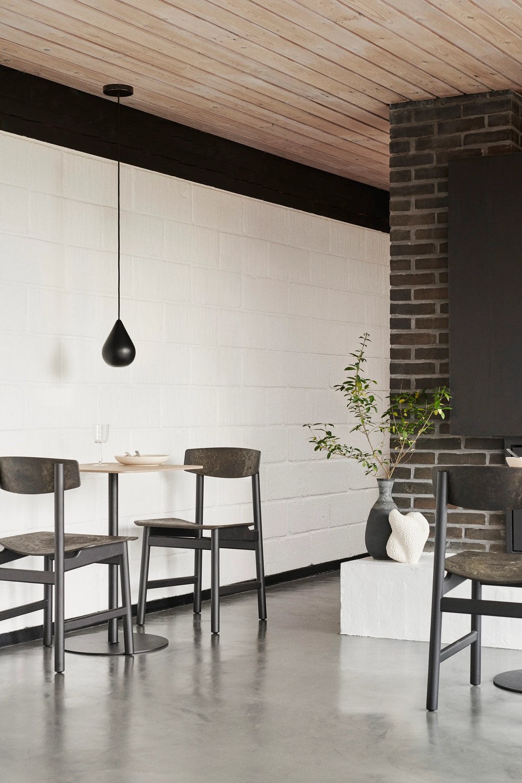 Two Mater Conscious Chairs in a dark, charcoal finish, paired with a small round dining table in a contemporary setting. The chairs feature a robust, eco-friendly design, complementing the industrial-chic interior with brick and white textured walls.