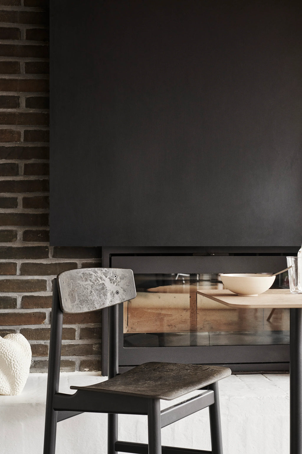 Mater Conscious Chair in a dark, charcoal finish, positioned beside a wooden table in a stylish dining area. The chair's textured seat and backrest are visible, set against a dark brick fireplace, emphasizing its contemporary and robust design.