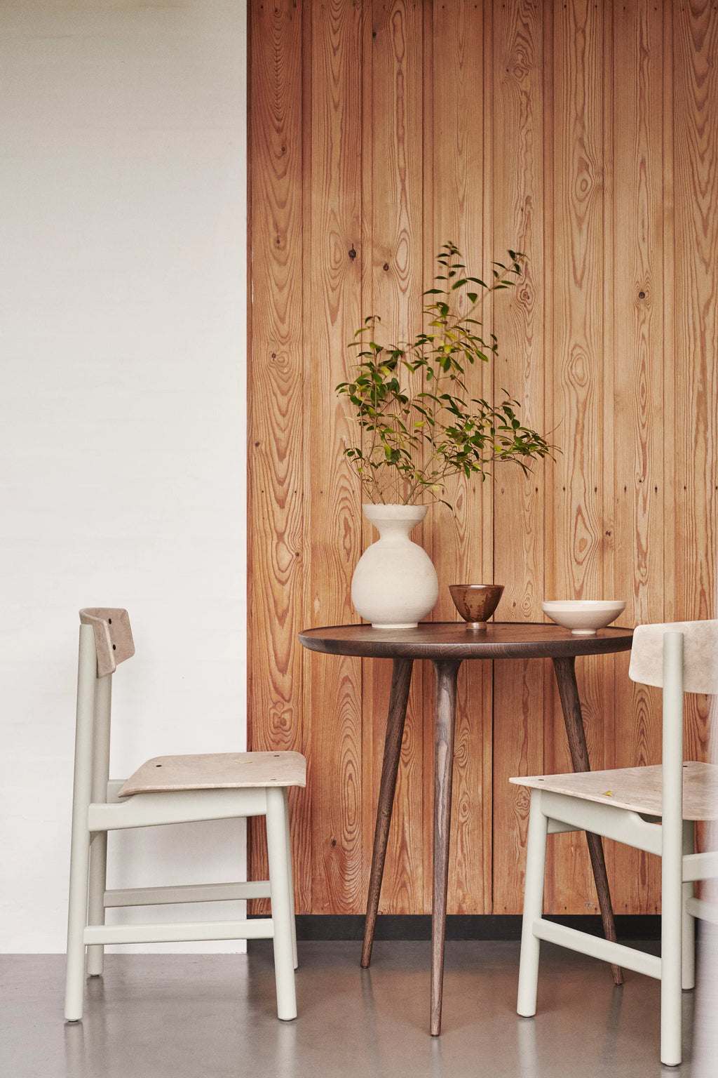 Two Mater Conscious Chairs in light, natural wood with a subtle pale green finish, flanking a dark wood round dining table. A white vase with green foliage adds a fresh, modern touch to the stylish interior.
