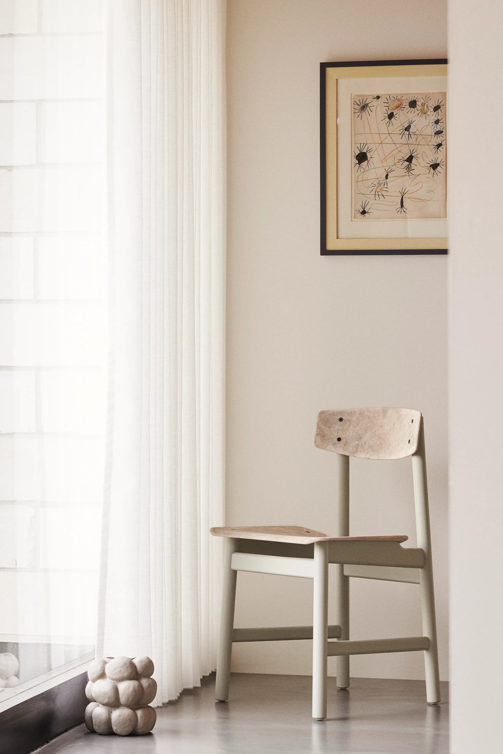 Mater Conscious Chair in a light green stained oak, with a natural recycled plastic seat and backrest, placed by a large window with sheer white curtains. The chair's gentle color and ergonomic design offer a calming presence in the room.