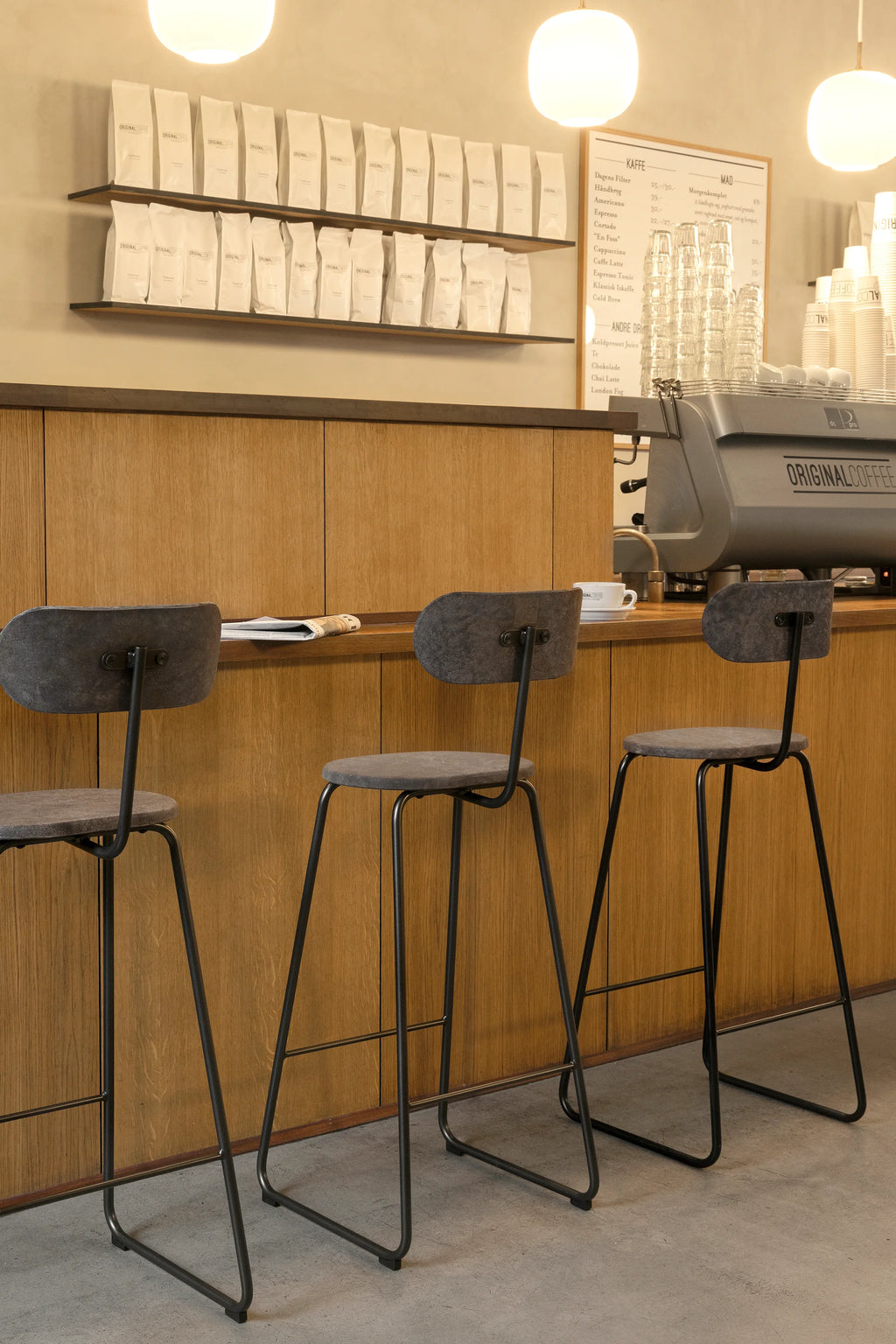 Mater Earth Counter Chair, a contemporary, sustainable design featuring a dark gray recycled composite seat and backrest, with elegant black metal legs, placed at a wooden counter in a cafe.