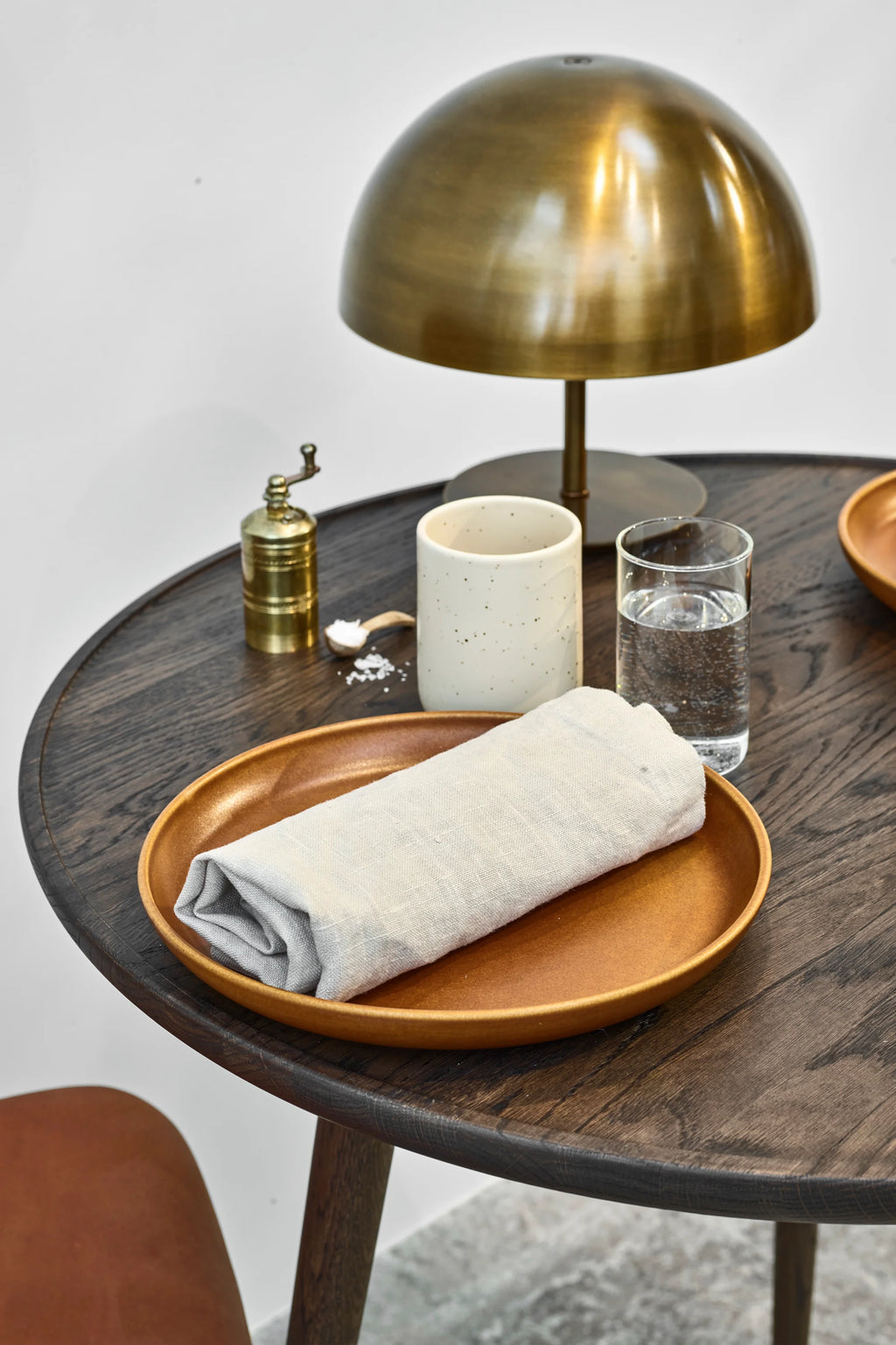 Mater Accent Round Dining Table, a dark stained oak finish, showcasing a detailed place setting with a golden plate, linen napkin, and brass lamp, emphasizing the table's elegant and functional design.