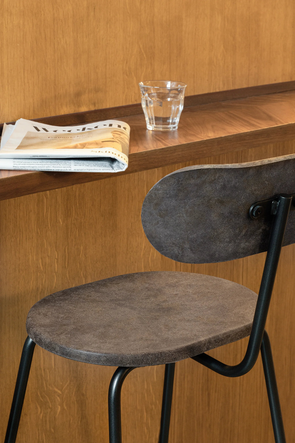Mater Earth Counter Chair, a modern dark gray textured counter stool with a black metal frame, shown at a warm wooden counter with a newspaper and glass of water.