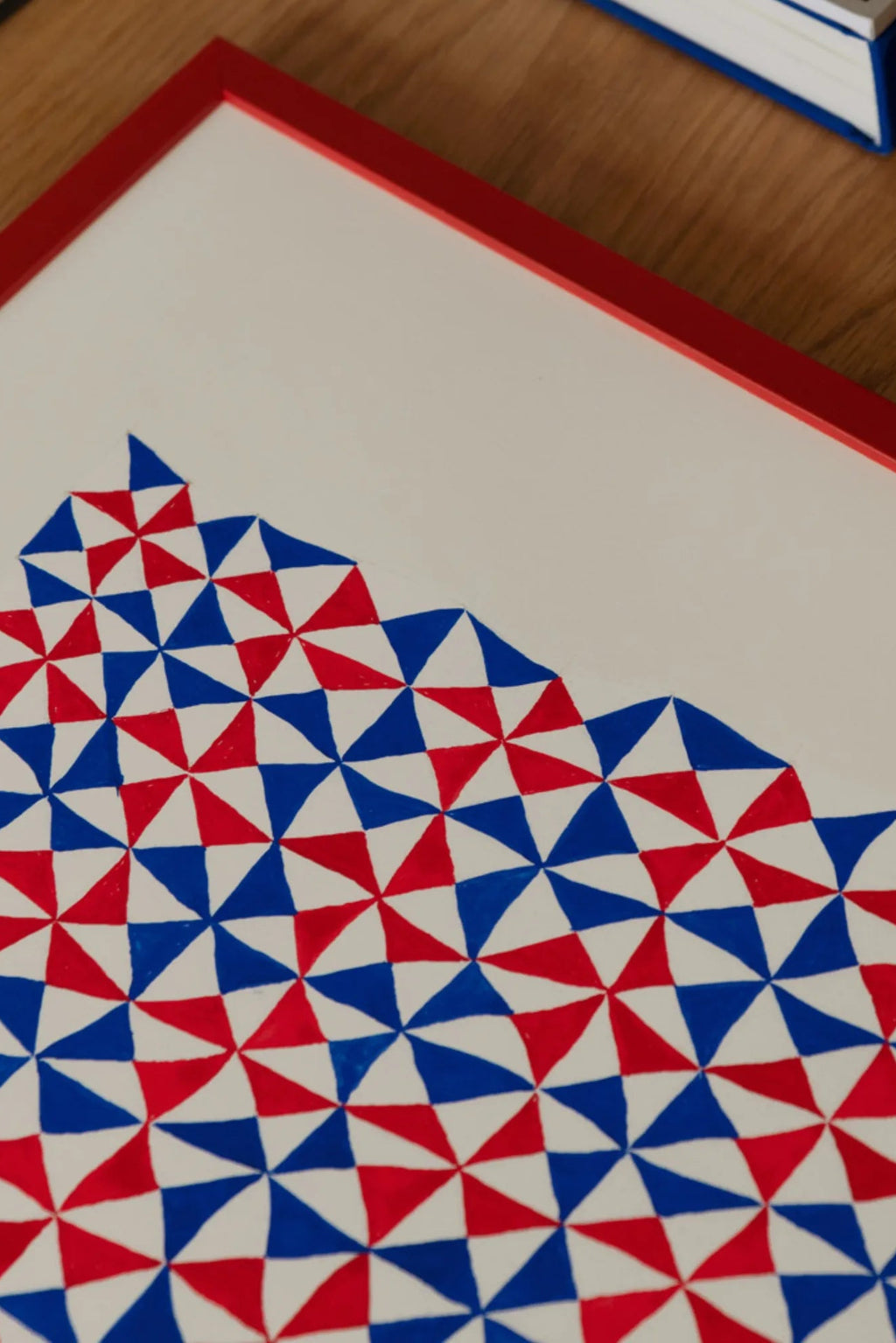 The Poster Club Tern No. 3, a vibrant geometric abstract art print featuring a dynamic pattern of interlocking red, blue, and white triangles, creating a visually striking optical effect. This graphic poster is presented within a bold red frame, adding a pop of color and definition. Ideal as modern wall art for contemporary living rooms, kids' rooms, or a playful addition to a gallery wall.