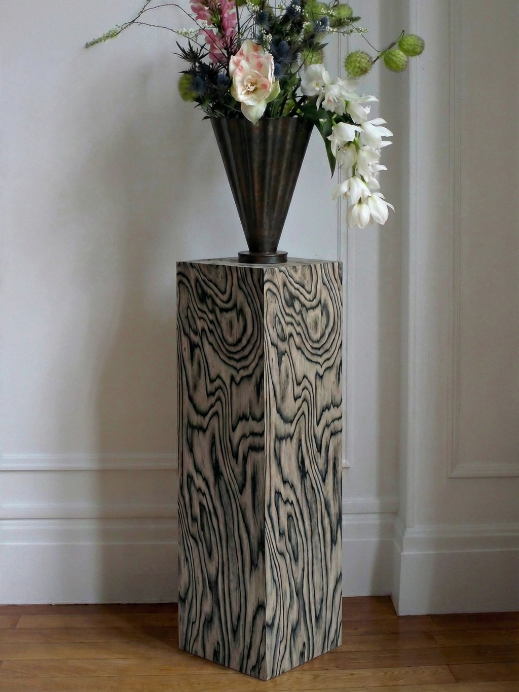 TCL Studios Monolith Sottsass Display Column, a tall, square postmodern pedestal table featuring a striking graphic wood grain veneer in contrasting light and dark tones. This sculptural side table, inspired by Memphis Group aesthetics, serves as an artistic plinth for showcasing decor or plants.
