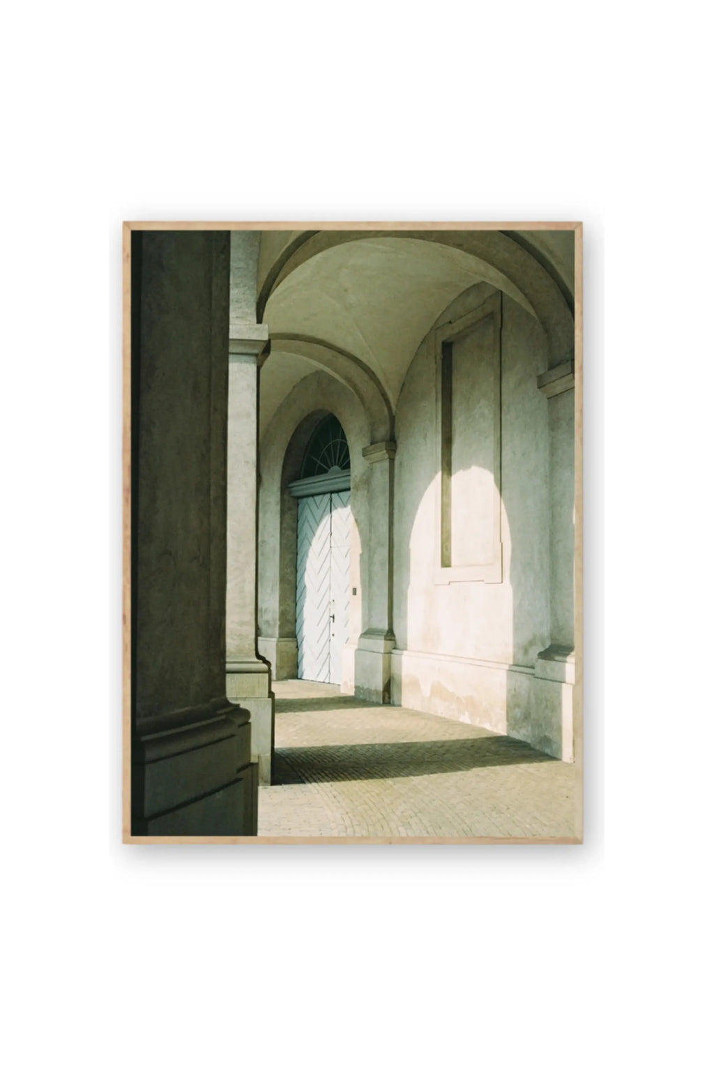 The Poster Club Morning Sun - Art Print, a premium photographic print capturing a sunlit arched hallway with light blue double doors and textured cream walls, framed in natural wood. This elegant architectural poster is perfect for modern home decor enthusiasts seeking stylish wall art.