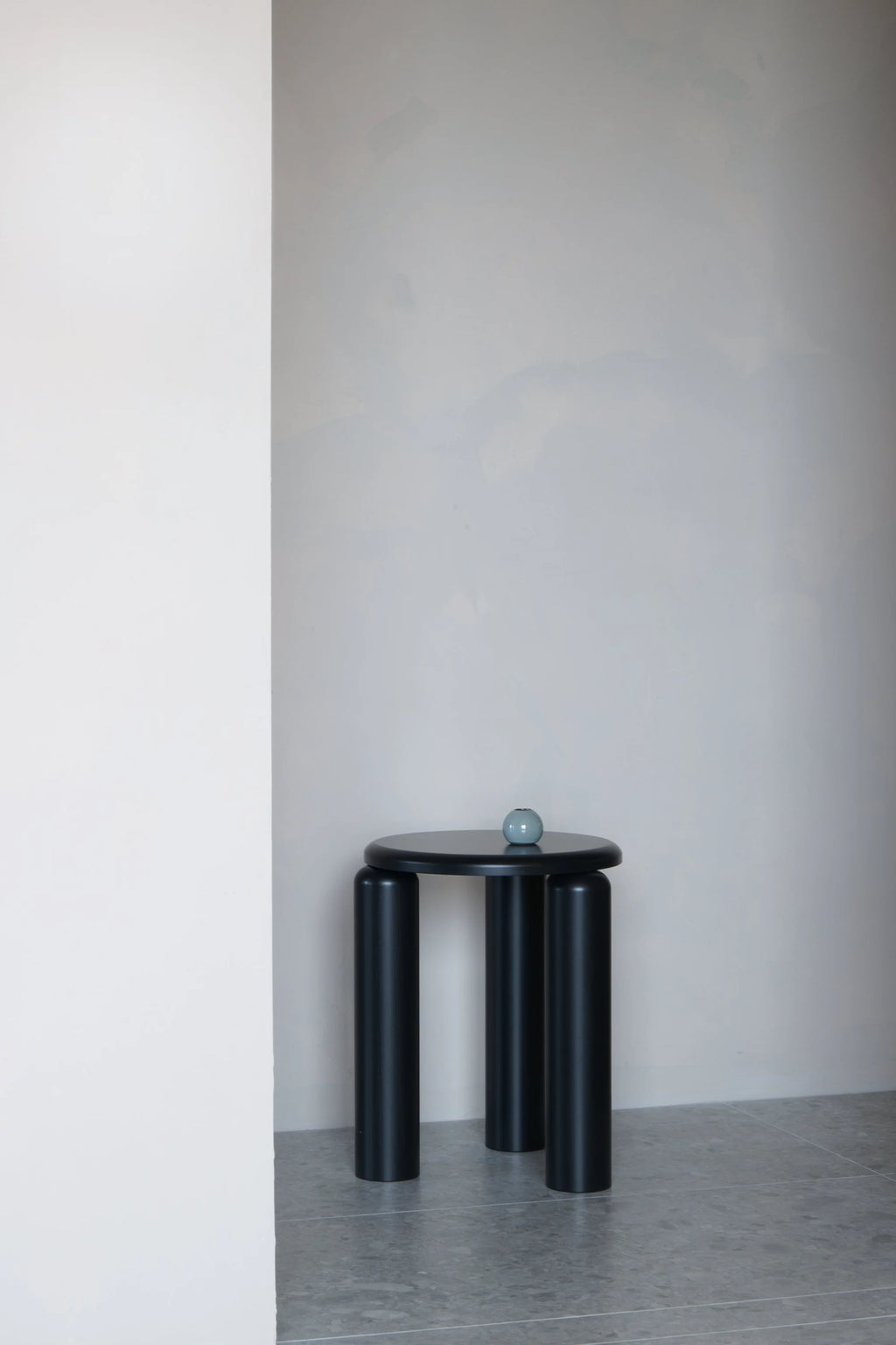 TAMO Neo Side Table in a minimalist, black matte finish, featuring a round top and three thick cylindrical legs on a tiled floor. A small teal vase sits on the top. Ideal for contemporary living spaces.