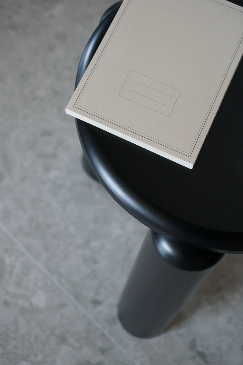 TAMO Neo Side Table close-up, featuring a minimalist black round top and a sturdy cylindrical leg, showcasing its modern design. A plain beige notebook rests on top. Ideal for contemporary living spaces.