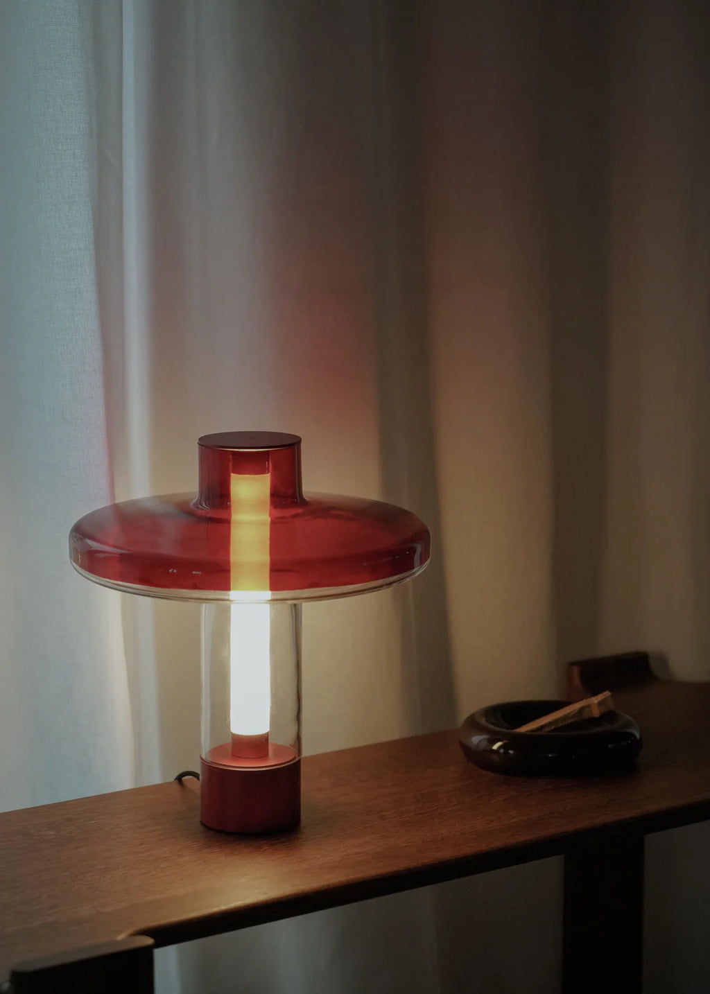 TAMO Nevo Table Lamp, a striking modern table lamp featuring a vibrant red blown glass disk shade and matching red base, with a clear cylindrical glass body housing a vertical light source. This contemporary design lamp casts a warm, inviting glow, perfect as a minimalist accent lamp for bedside tables, living room consoles, or stylish home office desks.
