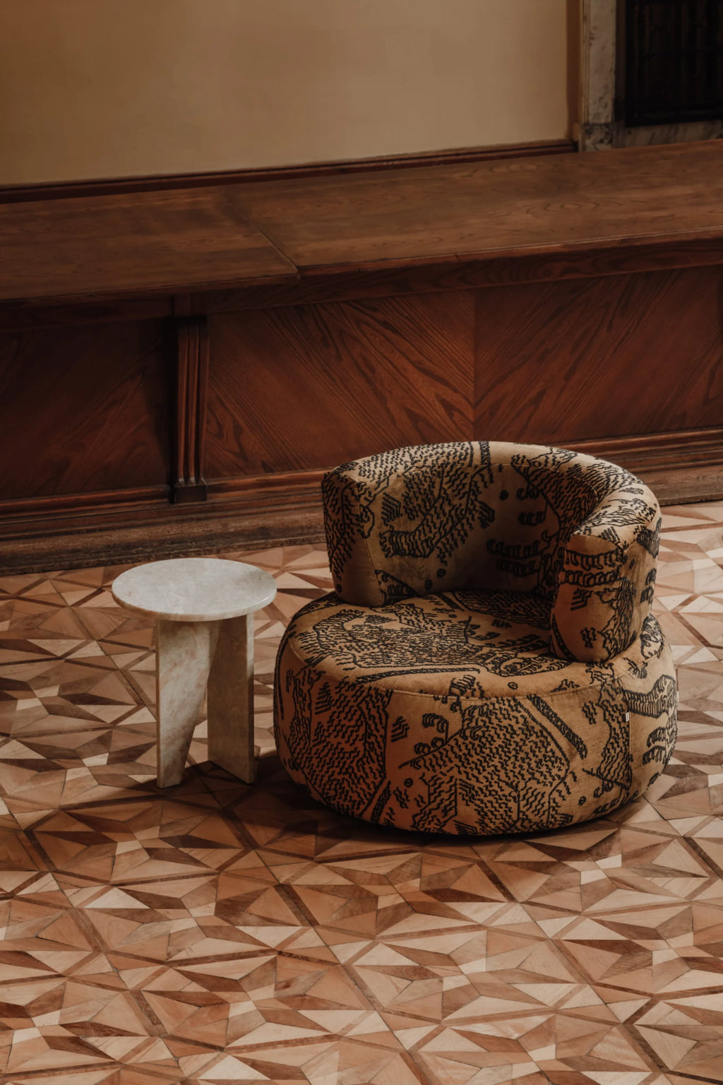 NG DESIGN Object 051 Lounge Chair, a premium upholstered club chair in a warm tan fabric with intricate black embroidered patterns, paired with a white marble side table on an ornate wooden floor. Ideal for luxury living spaces.