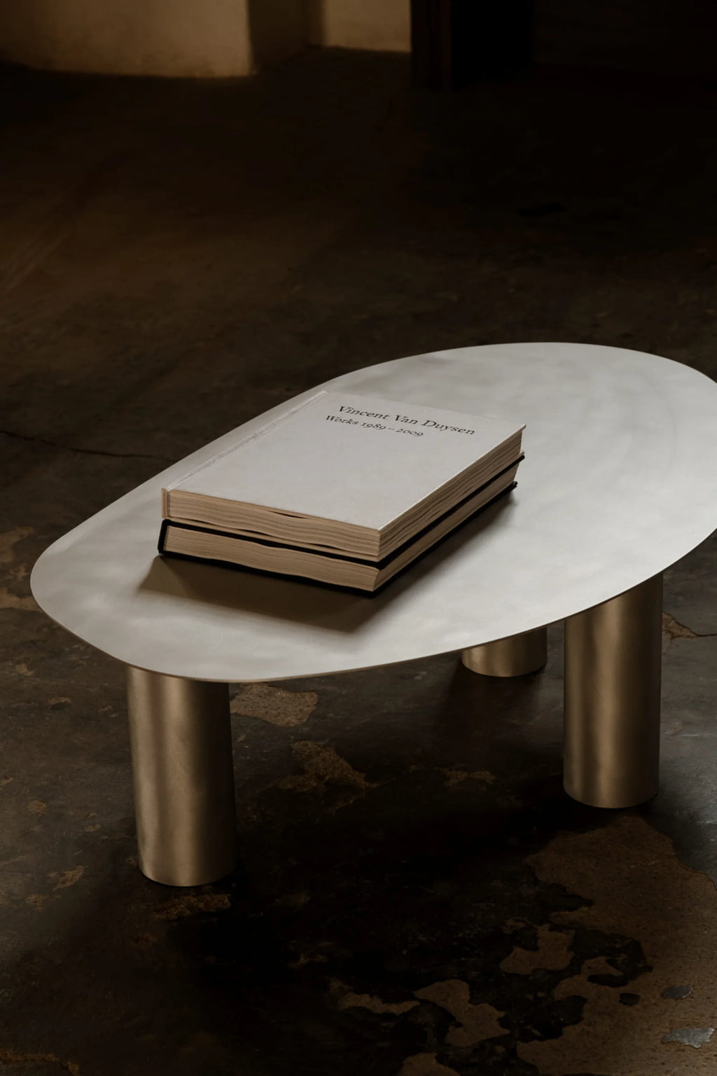 NG DESIGN Object 115 Coffee Table, a minimalist, oval-shaped brushed silver metallic table with three cylindrical legs, displaying a thick book on its surface. A luxury statement piece for modern living spaces.