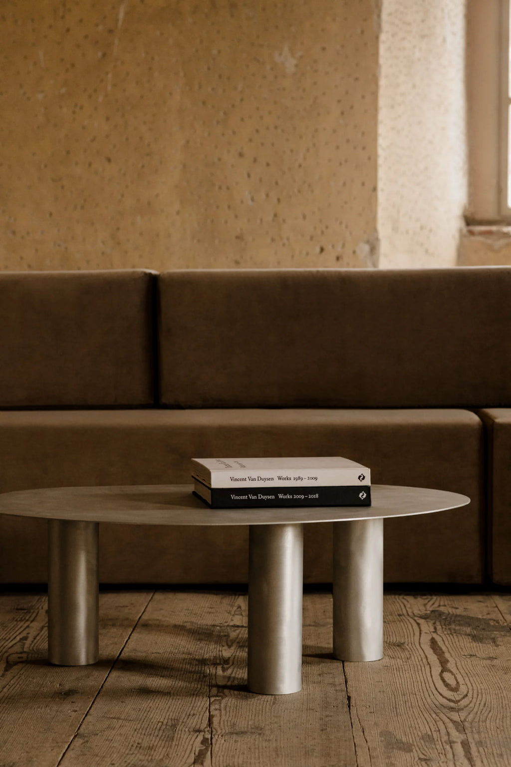 NG DESIGN Object 115 Coffee Table: A modern, minimalist low oval table with a matte silver finish and three thick cylindrical legs, displayed in a lifestyle setting on rustic wooden floorboards with a plush brown sofa in the background. An authentic design ideal for contemporary living spaces.