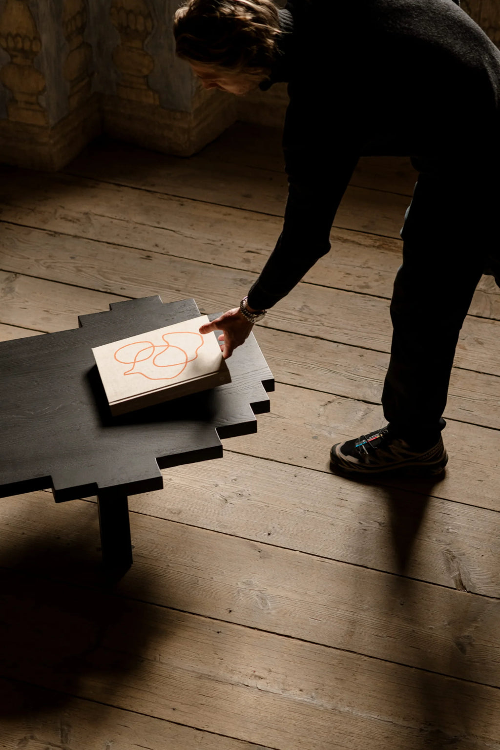 NG DESIGN Object 116 Coffee Table, a modern black wooden coffee table with a unique puzzle-piece-like top, seen from above with a person's hand placing a cream book on it. A perfect contemporary furniture accent.
