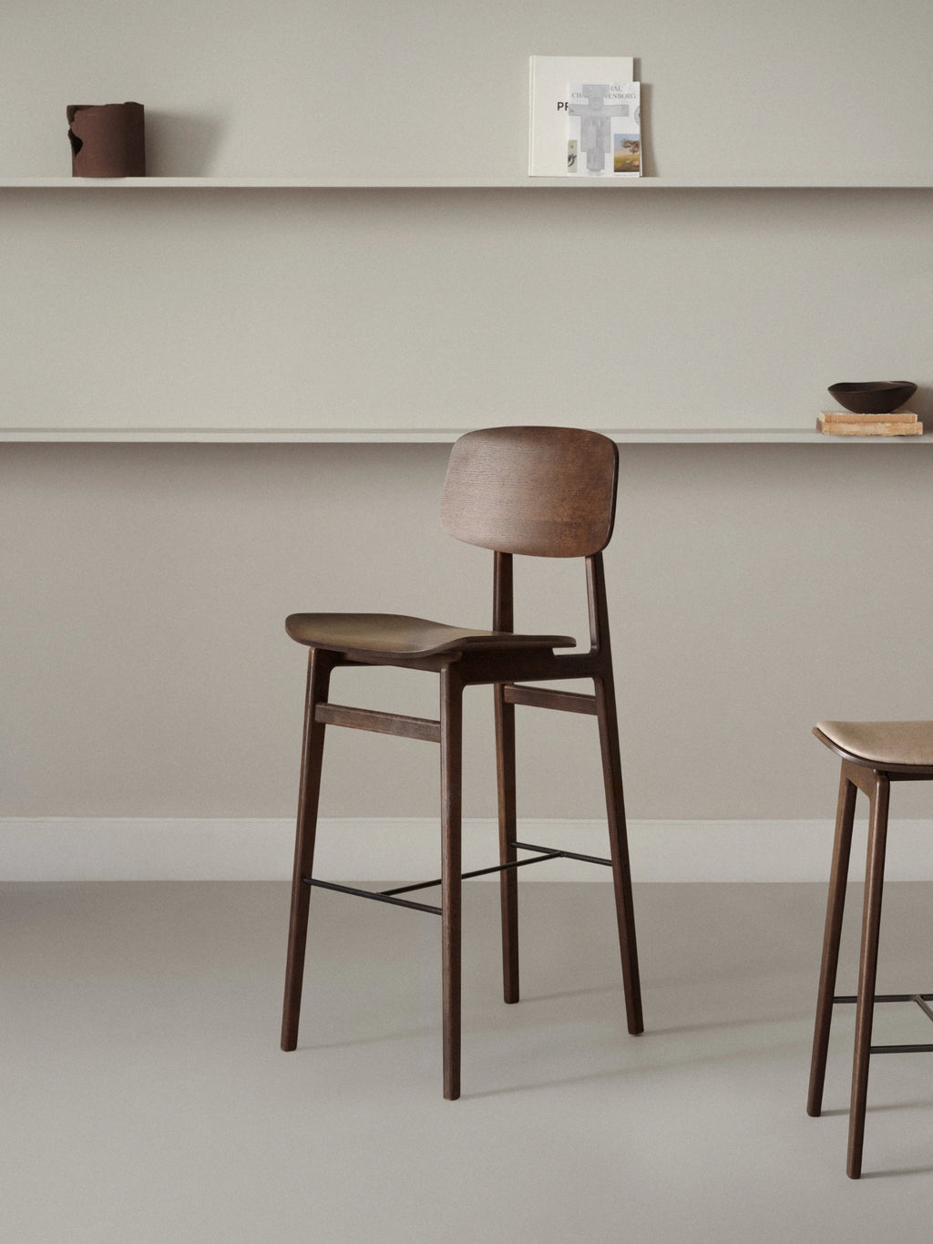 NORR11 NY11 Bar Chair, a minimalist Scandinavian bar stool crafted from dark stained solid oak wood with a gently curved backrest and seat for ergonomic comfort. This modern wood bar chair features a sleek silhouette with tapered legs and a matte black metal footrest. Ideal as a stylish kitchen counter stool or contemporary bar height seating for modern homes.