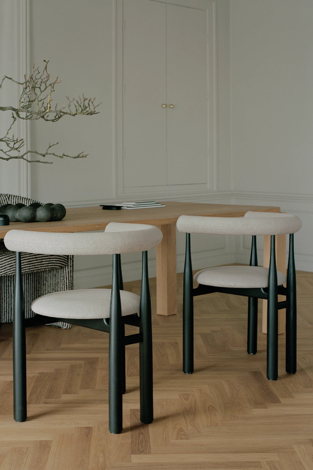 New Works Upholstered Bukowski Dining Chair, shown in a lifestyle shot with two chairs featuring light beige textured upholstery on black tapered legs. A premium design for modern dining.